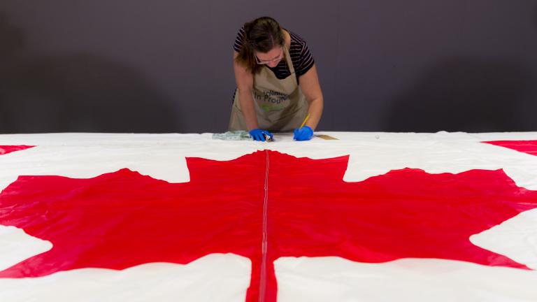 Une femme portant des gants de protection et un tablier prend des notes derrière un drapeau du Canada, posé à plat sur une table à l’avant-plan.