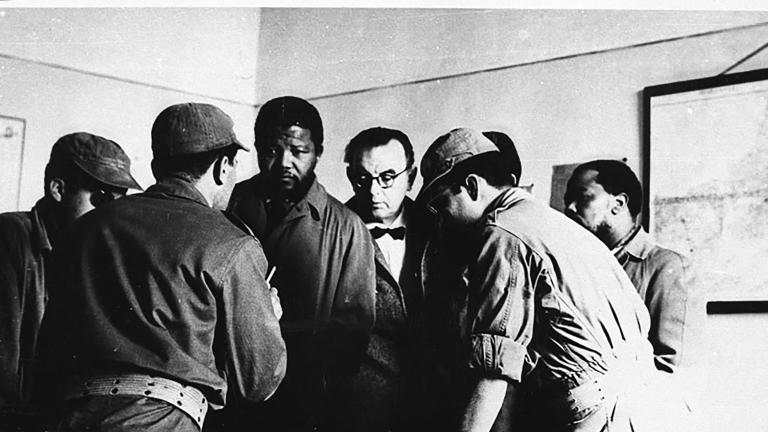 Une image en noir et blanc de Nelson Mandela debout devant un bureau. Il est entouré de cinq hommes, certains portant une tenue militaire. Un homme, le dos tourné à l’objectif, semble parler à Mandela, et les autres regardent quelque chose sur le bureau. 