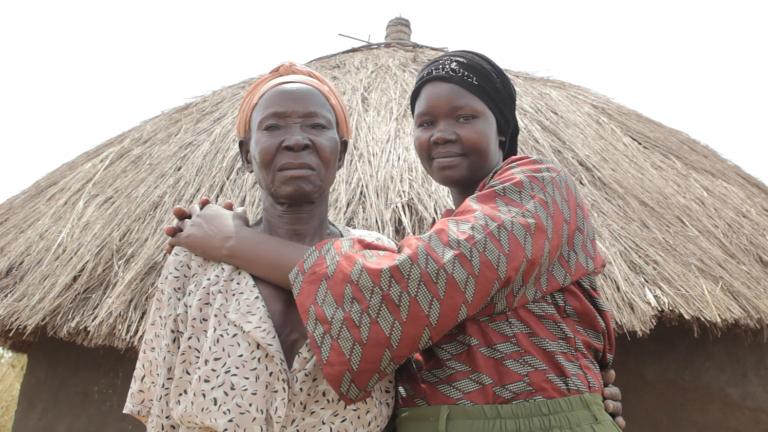 Deux femmes debout devant une hutte avec un toit de paille, regardent la caméra. La femme plus à droite sourit en serrant l’autre femme dans ses bras.