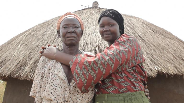Deux femmes debout devant une hutte avec un toit de paille, regardent la caméra. La femme plus à droite sourit en serrant l’autre femme dans ses bras. Visibilité masquée.