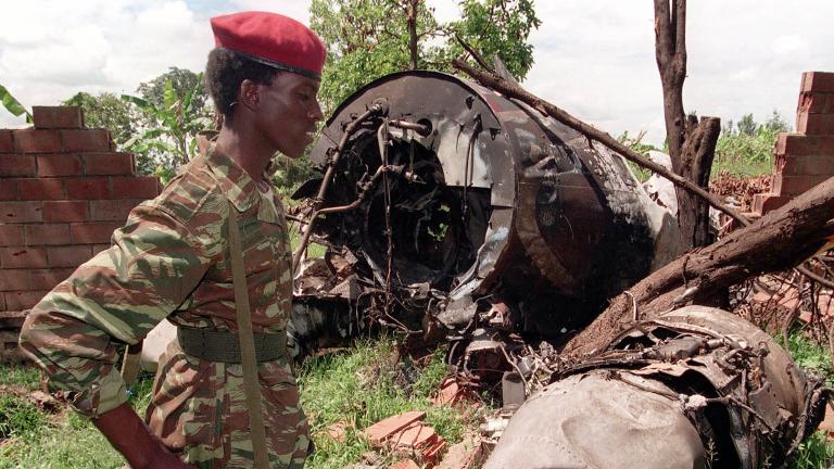 Un soldat portant un fusil, un béret rouge et un uniforme de camouflage se tient devant l’épave d’un avion au milieu des arbres.