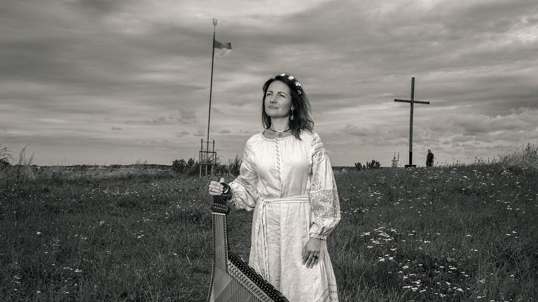 Une femme vêtue d'une longue robe blanche se tient dans un champ en tenant une bandura, un instrument folklorique ukrainien à cordes pincées. Visibilité masquée.