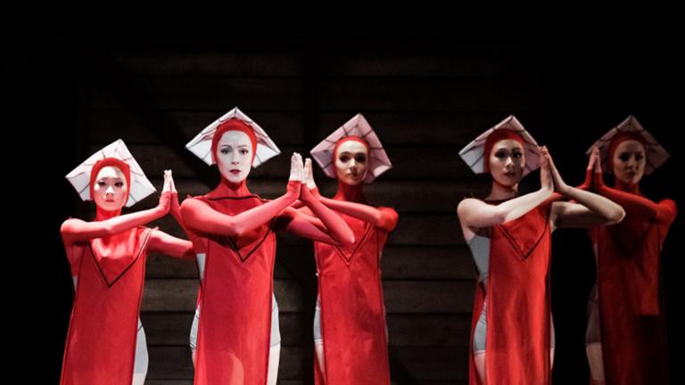 Sur une scène sombre se trouvent cinq danseuses de ballet vêtues de costumes rouges fluides aux genoux et coiffées de chapeaux carrés blancs. Elles regardent vers l'avant, les mains en prière. Visibilité masquée.