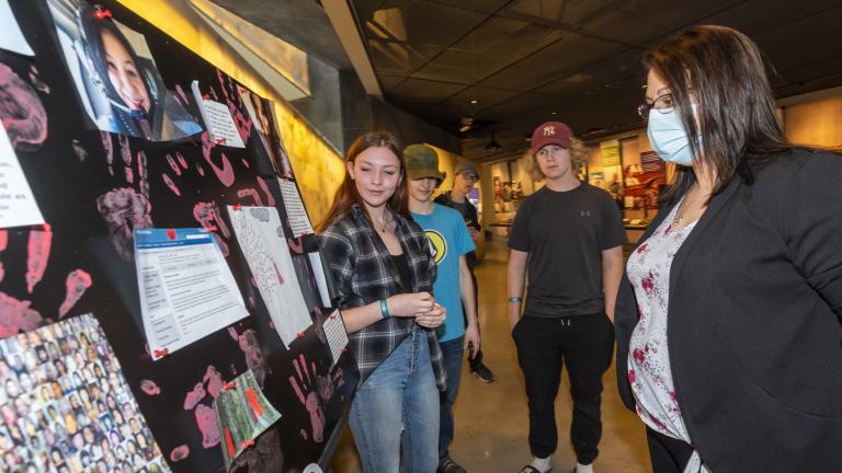 Trois élèves se tiennent devant un panneau d'affichage posé sur une table. Un visiteur du musée se tient devant eux et regarde le panneau d'affichage. Visibilité masquée.