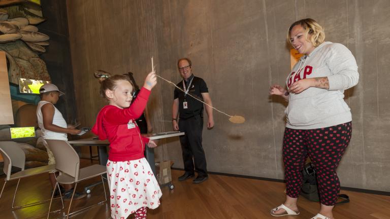 A young girl holds a small stick attached to a string and a small circular piece of cardboard. She is swinging this rope and cardboard as two adults watch. Visibilité masquée.