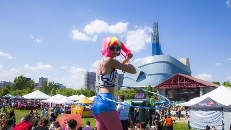 Une personne portant une perruque de couleur vive et utilisant un hula-hoop lors d’un festival en plein air; on voit le Musée dans l’arrière-plan. Visibilité masquée.