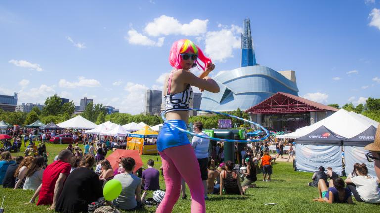 Une danseuse de hula hoop avec une perruque rose et jaune fluorescent et des leggings roses marche dans une foule de personnes assises sur un champ d'herbe devant un grand bâtiment en verre. Visibilité masquée.