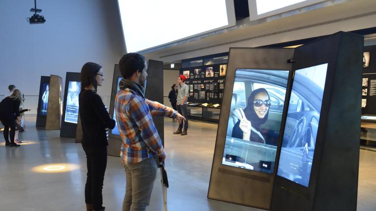 Un homme et une femme sont debout et regardent un écran vidéo en deux parties dans une galerie. L'homme a le bras allongé vers l'écran. Visibilité masquée.