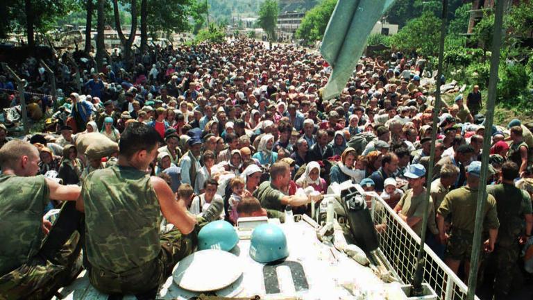 Deux soldats en uniforme sont assis sur un grand véhicule, surplombant une foule dense de personnes qui s’étend au loin. Visibilité masquée.