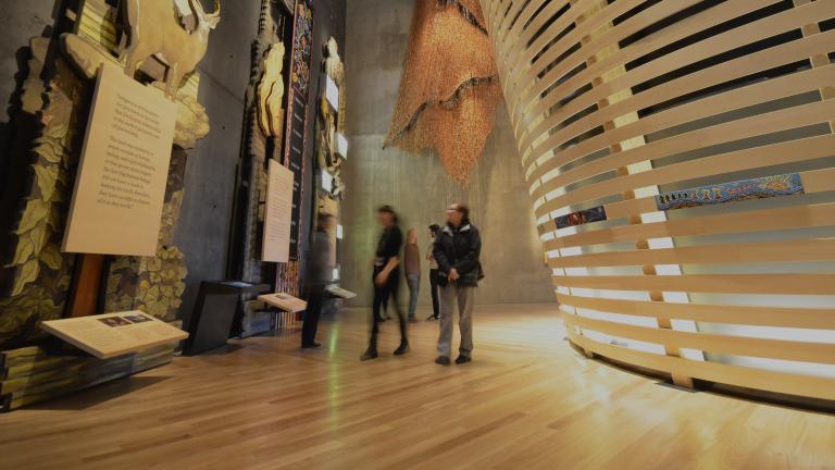 Quelques personnes dans une galerie du Musée explorent de grands panneaux de bois décorés de figures d’animaux et d’une œuvre perlée métisse. À droite, on voit des lattes de bois courbées placées horizontalement.