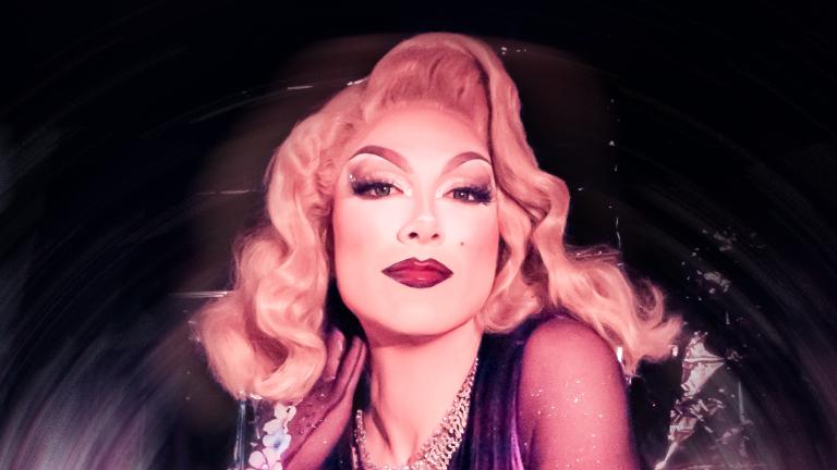 Une drag queen aux cheveux blonds mi-longs, portant des bijoux argentés et une robe violette, pose devant un fond argenté. Visibilité masquée.
