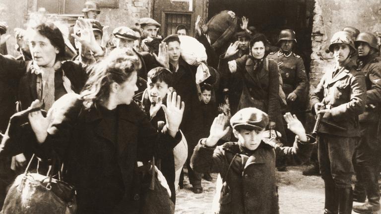 Photo en noir et blanc montrant un groupe de personnes les mains levées, sous la menace de soldats armés. Au premier plan, un jeune garçon effrayé.