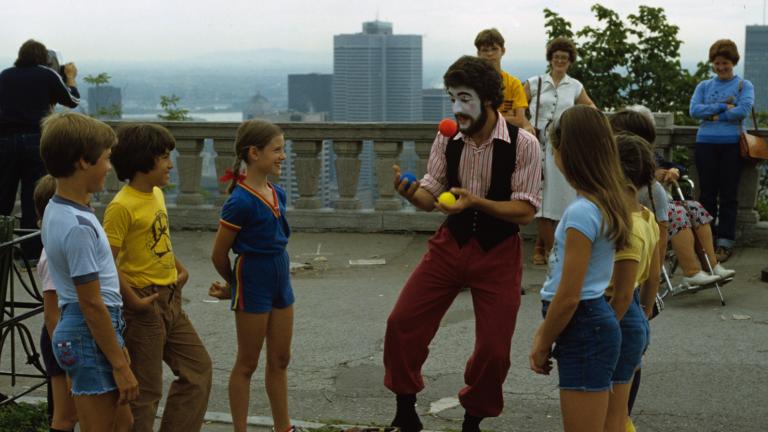 Un homme au visage peint jongle en plein air, sous le regard d'enfants souriants. Une silhouette de ville apparaît au loin derrière lui.