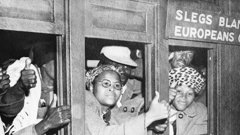 Une image en noir et blanc d’un groupe de femmes et d’hommes noirs à l’intérieur d’un wagon qui regardent à l’extérieur par les fenêtres. Plusieurs ont la main levée et tiennent le pouce en l’air. Au-dessus d’une fenêtre, il est écrit que le wagon est réservé à la population européenne.