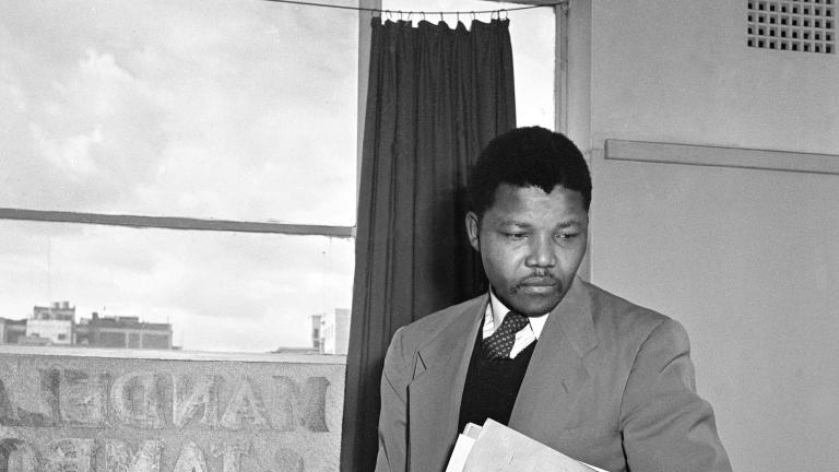 Une image en noir et blanc de Nelson Mandela, un homme noir vêtu d’un complet et portant un livre et quelques dossiers sous le bras gauche, debout derrière un bureau d’un cabinet.