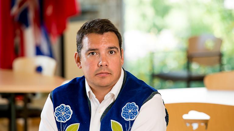 A man wearing a blue vest with flower beadwork, seated with his hands resting on a table. Visibilité masquée.