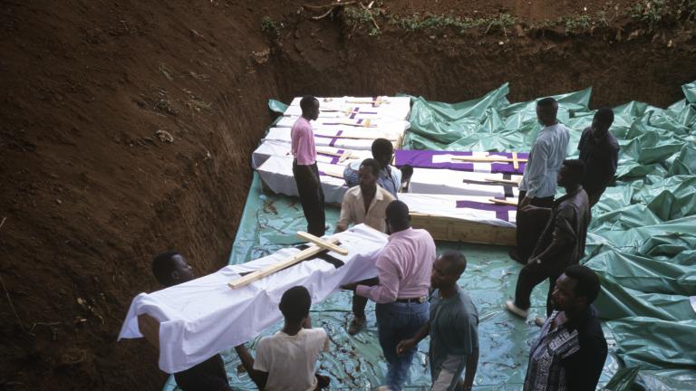 Plusieurs hommes portent un cercueil enveloppé de tissu et orné d’une grande croix en bois dans une grande fosse commune creusée dans la terre qui contient déjà plusieurs cercueils similaires.