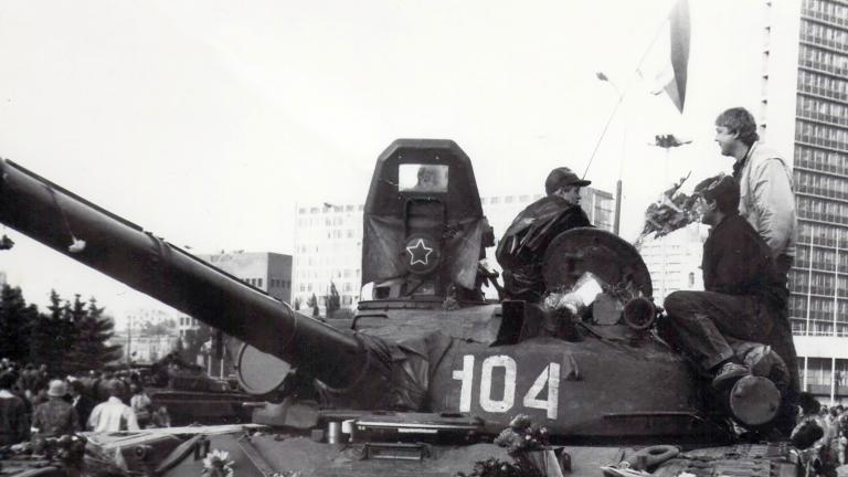 Deux hommes debout devant un char militaire stationné. Il y a des bouquets de fleurs sur le char. Derrière, on voit des gens déambuler devant un haut édifice. La photo est en noir et blanc.