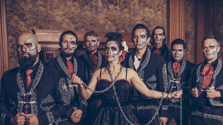 Les membres du The Mariachi Ghost, debout en rang, sont vêtus de costumes noirs assortis avec des détails cousus blancs et des cravates rouges. La moitié gauche de leur visage est peinte pour ressembler à un squelette. Visibilité masquée.
