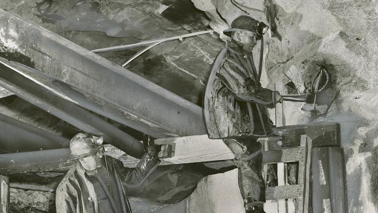 Deux mineurs portant un casque travaillent sous terre dans une mine d'uranium. L'un est debout sur une échelle alors que l'autre le regarde.
