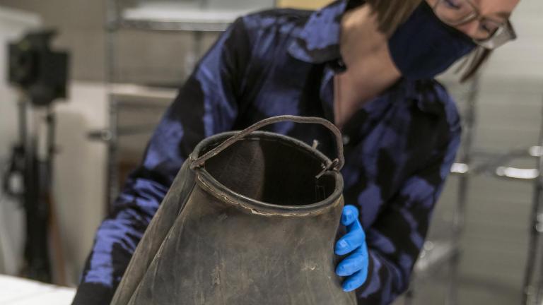 Une femme tient un sac en caoutchouc noir éraflé avec une poignée métallique courbée de forme grossière sur le dessus.