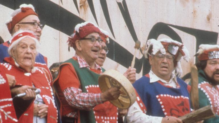 Un groupe d’hommes et de femmes plus âgés chante, debout devant un grand bâtiment en bois. Un homme joue du tambour. Tous et toutes portent de longues chemises ou des couvertures décorées de motifs faits de boutons.