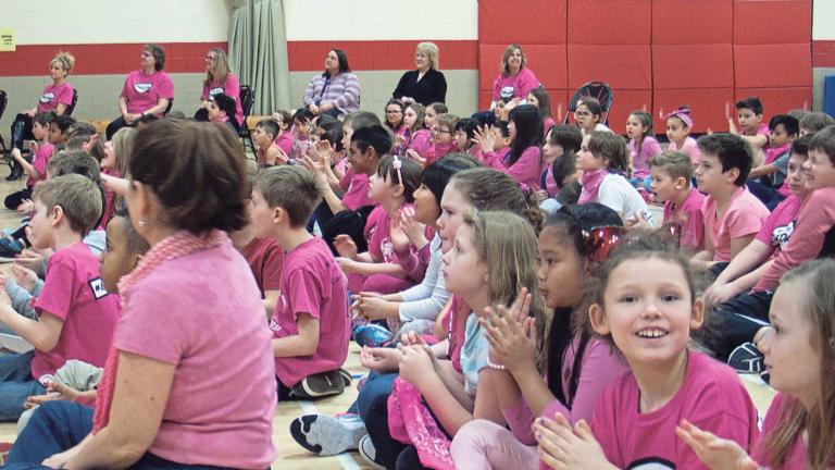 Un grand groupe d'enfants sont assis dans un gymnase d'école. Ils portent tous des chandails ou des foulards roses. Ils sont tous attentifs à quelque chose qui est hors cadre, sauf une fille qui sourit vers l'objectif.