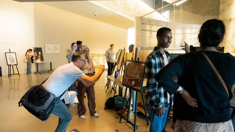 Des projets d’élèves sont exposés sur des chevalets dans une galerie de musée. Deux élèves parlent de leur travail avec des adultes. Visibilité masquée.