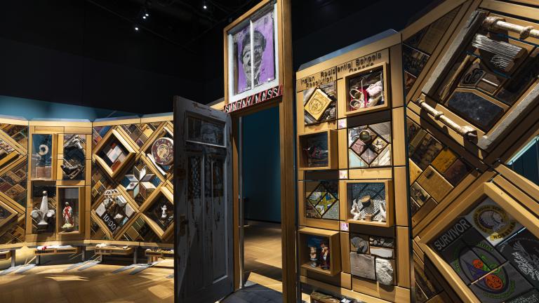 Un grand mur de bois incurvé et autoportant sur lequel sont fixés de nombreux petits objets disposés en losanges et en rectangles. Il y a une porte entrouverte au centre du mur. Visibilité masquée.