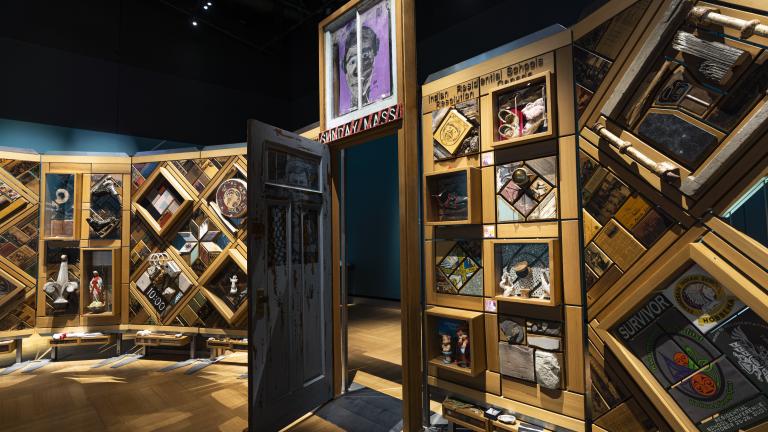 Un grand mur de bois incurvé et autoportant sur lequel sont fixés de nombreux petits objets disposés en losanges et en rectangles. Il y a une porte entrouverte au centre du mur. Visibilité masquée.