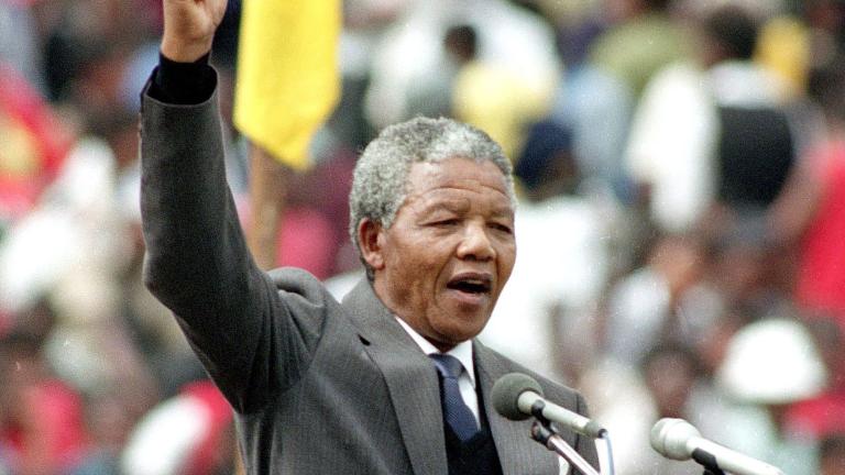 Nelson Mandela, un homme plus âgé à la peau foncée et aux cheveux gris et courts, est debout sur un podium et parle dans deux microphones. Il porte un complet gris et une cravate. Il lève son poing droit au-dessus de sa tête. Visibilité masquée.