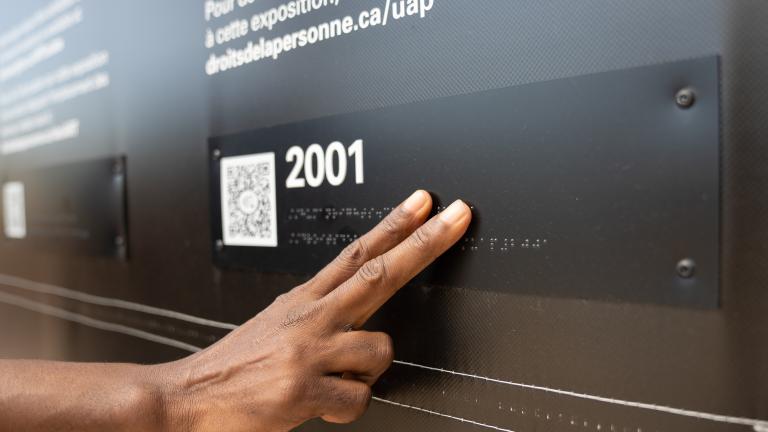 Une main touche des lettres en braille dans une exposition muséale. Visibilité masquée.