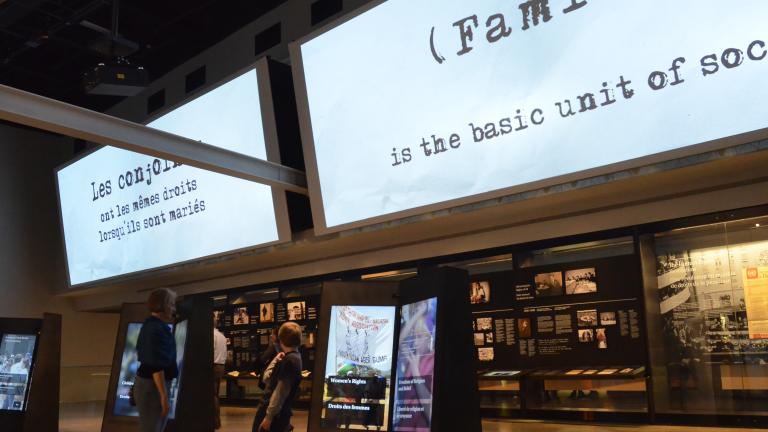 Une femme et un enfant interagissent avec un élément d'exposition numérique dans une galerie de musée. Au-dessus d'eux, sur un grand écran, on peut lire, en anglais, "La famille est l'unité de base de la société." Visibilité masquée.