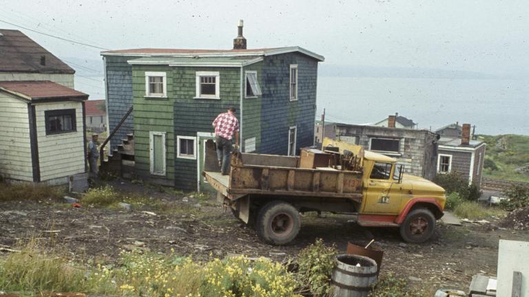 Un homme se tient à l’arrière d’un camion à ordures garé devant une maison en bois aux bardeaux verts. À l’arrière-plan, on aperçoit des rails de chemin de fer et d’autres maisons, et plus loin encore, le port d’Halifax.