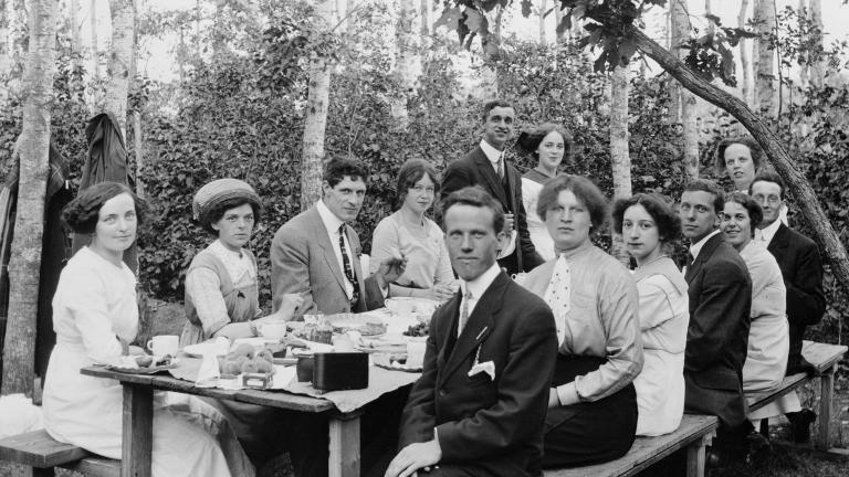 Un groupe d’hommes et de femmes bien habillés assis à une table de pique-nique avec des arbres en arrière-plan. 