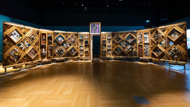 Un grand mur de bois incurvé et autoportant sur lequel sont fixés de nombreux petits objets disposés en losanges et en rectangles. Il y a une porte entrouverte au centre du mur. Visibilité masquée.