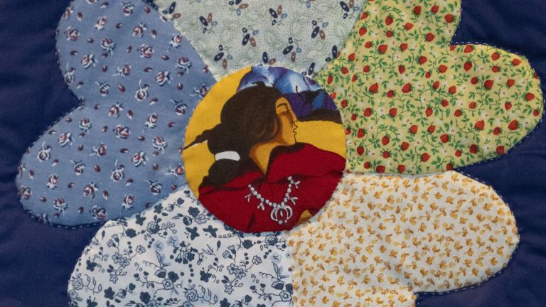 Gros plan sur une fleur sur une courtepointe. Chacun des pétales est un tissu fleuri différent, tandis qu’au centre de la fleur se trouve un tissu sur lequel est imprimé le profil d’une femme autochtone. Visibilité masquée.