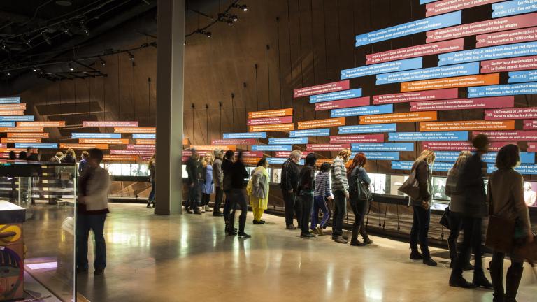 Une grande foule dans une galerie du Musée. La plupart des gens regardent une exposition sur le mur de la galerie. L'exposition comprend une série d'images et de longs panneaux rectangulaires de texte, placés horizontalement. Visibilité masquée.