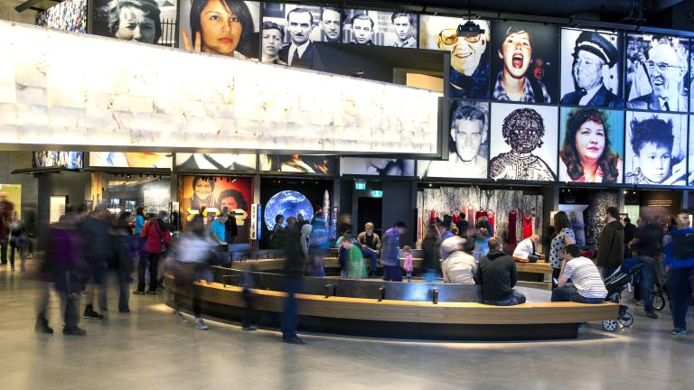 Une grande foule dans une galerie de musée. Il y a un banc courbe au centre de la salle. Dans l’espace, on voit une série de grandes images placées au-dessus d’alcôves qui contiennent chacune une exposition différente.