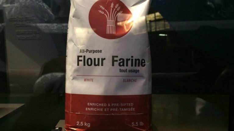 Un sac de farine dans une exposition. Le texte sur le sac lit comme suit : All-Purpose Flour/Farine tout usage, White/Blanche, Enriched & Pre-Sifted/Enrichie et pré-tamisée, 2.5 kg/5.5lb