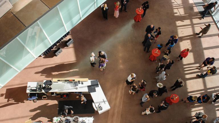 Une vue à vol d'oiseau d'une foule de personnes rassemblées dans un grand espace ouvert du Musée. Il y a quatre tables de cocktail recouvertes de nappes rouges et un bar sur le côté gauche de l'image. Visibilité masquée.
