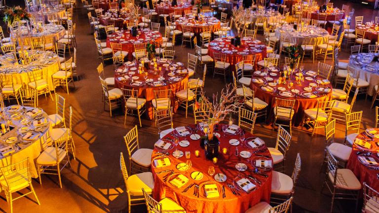 Plusieurs tables rondes, chacune avec des couverts formels pour 10 personnes, vues d'en haut. Une lueur orange est diffusée dans l’espace.