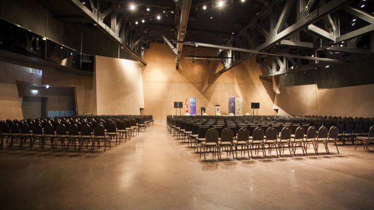 Une grande salle avec de hauts plafonds et un sol en béton brun poli. Quelques centaines de chaises sont alignées en rangs bien ordonnés face à une scène.