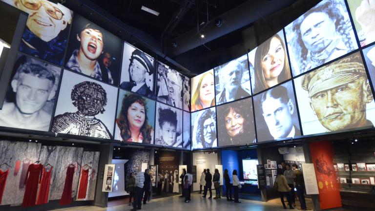 Des personnes explorent une galerie du Musée avec deux rangées de portraits carrés au-dessus d'une série d'alcôves, contenant des photographies, des vidéos et des panneaux de texte. Visibilité masquée.