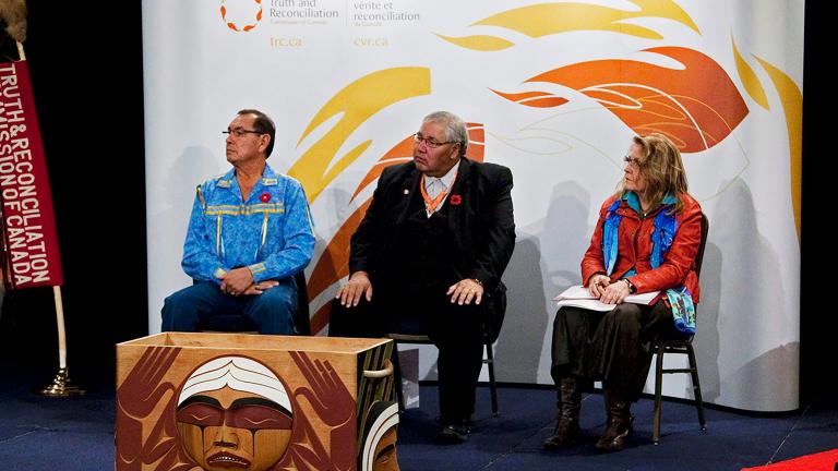 Deux hommes et une femme assis devant un arrière-plan blanc décoré de flammes stylisées. À leur droite, on voit une bannière verticale sur laquelle on peut lire « Truth & Reconciliation Commission of Canada ». et devant eux se trouve une boîte rectangulaire en bois sculpté. Sur le côté qui fait face à l'objectif, on voit un visage rond qui pleure.