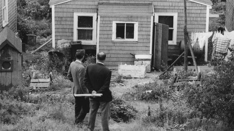 Une image en noir et blanc de deux hommes en complet debout devant une maison en bois. Ils font dos à l’objectif.