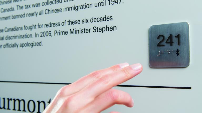 Une main s’avance vers un point d’accès universel, qui est une petite plaque de métal carrée fixée au mur. La plaque porte le numéro 241 en relief et en braille, de même que le symbole Bluetooth.