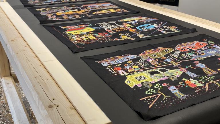 Une série de panneaux de broderie colorés sur du tissu noir, exposés sur une table de convoyeur en bois à manivelle et sur le mur d’un musée.