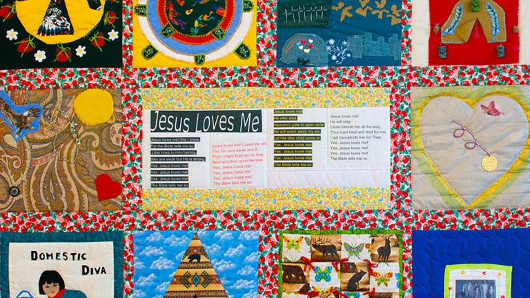 Une courtepointe colorée avec divers panneaux de tissu où figurent des images comme un oiseau, un cœur, un tipi, une femme nettoyant un plancher, et des lignes de texte sous le titre « Jesus Loves Me ».