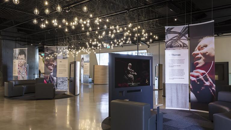 Une exposition de musée composée de divans courbes, d’écrans vidéo et de photos plus grandes que nature de personnes diverses.
